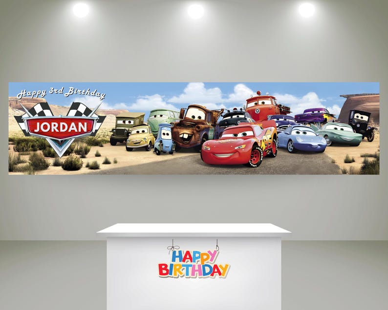 May include: A rectangular banner featuring a desert landscape with various animated car characters, including a red race car. The text reads Happy 3rd Birthday JORDAN. Below, a white table displays a colorful Happy Birthday sign in block letters.