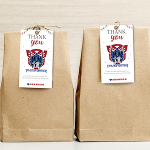 Könnte beinhalten: Zwei braune Papiertüten mit angehängten Dankeschön-Etiketten. Die Etiketten zeigen ein blaues und rotes Transformers-Logo mit dem Text "Danke" und "Transformer". Der Text "Ich fand den Kuchen und die Geschenke auch toll, aber mein Lieblingsteil war es, dich zu sehen!" und "Brandon" stehen ebenfalls auf den Etiketten.