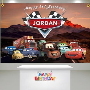 May include: A birthday banner featuring the Cars movie characters with the name "Jordan" in a red emblem. The banner has a mountain backdrop and the text "Happy 3rd Birthday". A smaller sign below reads "Happy Birthday".