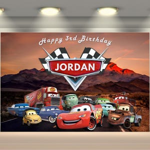 Puede incluir: Un tel&oacute;n de fondo con los personajes animados de Cars con una escena de monta&ntilde;a des&eacute;rtica. La pancarta dice "Happy 3rd Birthday JORDAN" con un emblema de bandera de carreras. La imagen incluye a Rayo McQueen y otros veh&iacute;culos.