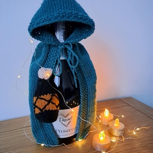May include: A teal knitted bottle cover, shaped like a cloak with a hood, encloses a Prosecco bottle. A small, black and orange knitted pouch hangs from the cover. The bottle is surrounded by small, lit candles and fairy lights.