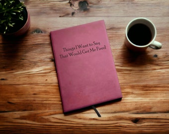 May include: A burgundy notebook with the text "Things I Want to Say That Would Get Me Fired" on the cover. The notebook is on a wooden table with a cup of coffee.
