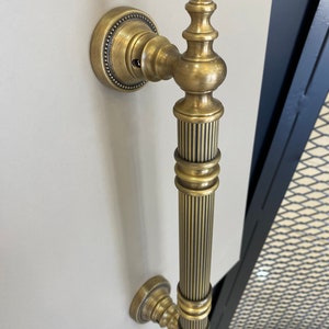 May include: A brass door handle with a fluted design and a decorative top. The handle is mounted on a white wall.