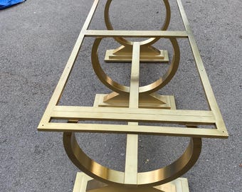 May include: A gold metal table base with a unique curved design. The base has three legs and is made of a sturdy metal.