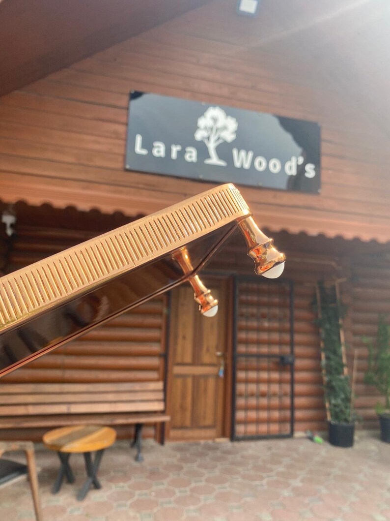 K&ouml;nnte beinhalten: Ein goldfarbenes Tablett aus Metall mit einem dekorativen, erh&ouml;hten Rand und drei Beinen. Das Tablett befindet sich vor einem Holzgeb&auml;ude mit einem Schild, auf dem "Lara Wood's" steht.