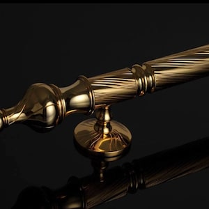 May include: A polished gold-colored door handle with a decorative design. The handle features a detailed, swirling pattern and a rounded end. The handle is mounted on a circular base, reflecting the light.