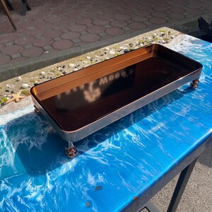 K&ouml;nnte beinhalten: Ein rechteckiges Tablett mit einer dunkelbraunen Oberfl&auml;che und einem goldenen Rand steht auf vier kleinen goldenen Beinen. Das Tablett befindet sich auf einer blauen und wei&szlig;en Oberfl&auml;che, die einer Strandszene &auml;hnelt.
