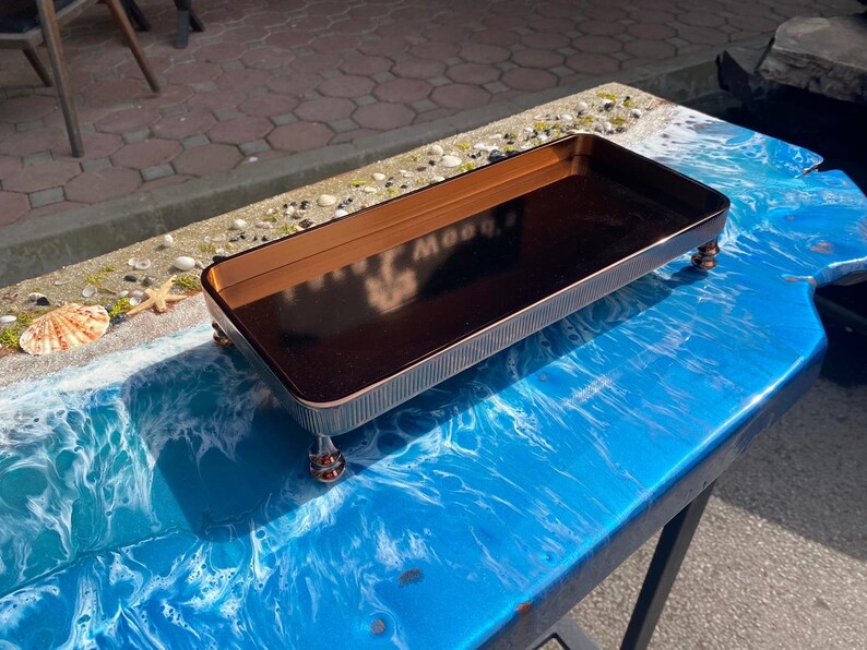 K&ouml;nnte beinhalten: Ein rechteckiges Tablett mit einer dunkelbraunen Oberfl&auml;che und einem goldenen Rand steht auf vier kleinen goldenen Beinen. Das Tablett befindet sich auf einer blauen und wei&szlig;en Oberfl&auml;che, die einer Strandszene &auml;hnelt.