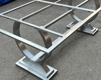 May include: A silver metal table frame with a rectangular top and curved legs. The table frame is sitting on a gray asphalt surface.