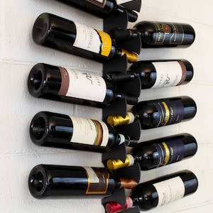 Peut inclure: Un porte-bouteilles mural en métal noir avec huit bouteilles de vin. Les bouteilles sont disposées en quinconce, les étiquettes étant tournées vers l'extérieur. Le porte-bouteilles est monté sur un mur en briques blanches.