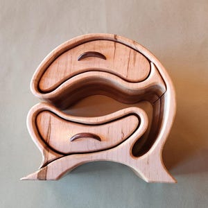 Puede incluir: Una joyero de madera hecho a mano con un diseño único y fluido. La caja presenta tres cajones con frentes curvos y pequeños pomos de madera marrón oscuro. La madera de color claro tiene patrones de vetas naturales.