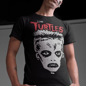 May include: A black t-shirt featuring a white graphic of a person's face with skull makeup. The text "I LIKE TURTLES" is printed in red above the graphic.