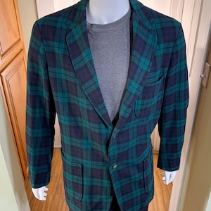 May include: A blazer featuring a dark green and navy blue plaid pattern with a single gold-toned button closure, notched lapels, two lower patch pockets, and one chest patch pocket, worn over a grey textured long-sleeve shirt.