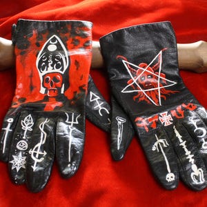 May include: A pair of black leather gloves featuring hand-painted designs. The left glove displays a red skull wearing a mitre, while the right glove features a red goat head inside a white pentagram. Both gloves are decorated with white symbols.