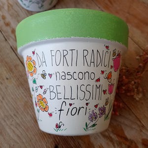 Può includere: Vaso di fiori in ceramica bianca con bordo verde, dipinto a mano con fiori colorati, api e cuori. Il testo italiano "DA FORTI RADICI NASCONO BELLISSIMI fiori" è scritto sul vaso. Un secondo vaso con fiori è visibile sullo sfondo.