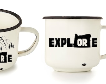 May include: Two white enamel mugs with black rims. One mug features a black silhouette of mountains and trees with the text "EXPLORE" and the state of Oregon. The other mug features the text "EXPLORE" with the state of Oregon.