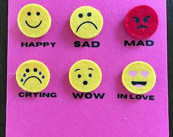 May include: A pink felt board with six emoji faces representing different emotions. The emojis are yellow, red, and pink, with text labels including "HAPPY," "SAD," "MAD," "CRYING," "WOW," and "IN LOVE."