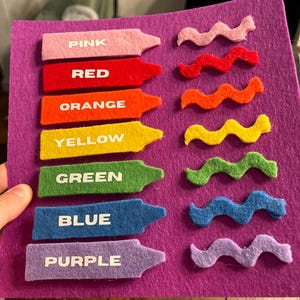 May include: A felt color learning set featuring seven crayon-shaped pieces labeled with colors: pink, red, orange, yellow, green, blue, and purple. Each crayon has a matching wavy felt piece. The set is on a purple felt background.