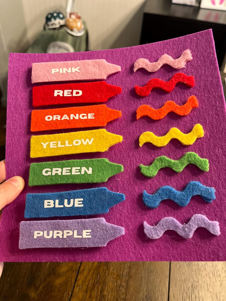 May include: A felt color learning set featuring seven crayon-shaped pieces labeled with colors: pink, red, orange, yellow, green, blue, and purple. Each crayon has a matching wavy felt piece. The set is on a purple felt background.