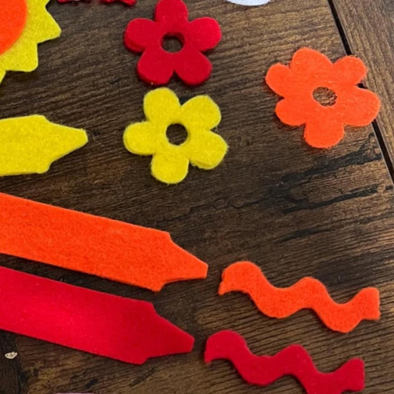 May include: A collection of colorful felt shapes, including flowers, crayons, and wavy lines, in shades of red, orange, and yellow. The shapes are arranged on a dark wooden surface, creating a playful and artistic composition.