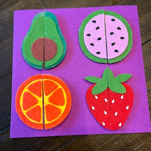 May include: A purple felt board features four felt fruit puzzles: an avocado, watermelon, orange, and strawberry. Each fruit is cut in half, creating a puzzle. The avocado is green and brown, the watermelon is pink and green, the orange is orange and yellow, and the strawberry is red and green.
