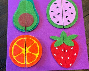 May include: A purple felt board features four felt fruit puzzles: an avocado, watermelon, orange, and strawberry. Each fruit is cut in half, creating a puzzle. The avocado is green and brown, the watermelon is pink and green, the orange is orange and yellow, and the strawberry is red and green.