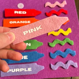 May include: A felt color matching activity with a purple background. The image shows felt crayons in red, orange, pink, green, blue, and purple, each labeled with its color name. Wavy felt shapes in various colors are also present.