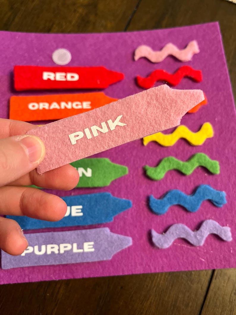May include: A felt color matching activity with a purple background. The image shows felt crayons in red, orange, pink, green, blue, and purple, each labeled with its color name. Wavy felt shapes in various colors are also present.