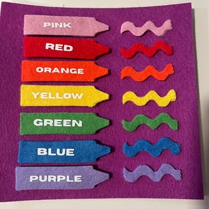May include: A purple felt board displays color learning tools. Seven crayon-shaped felt pieces, labeled "PINK," "RED," "ORANGE," "YELLOW," "GREEN," "BLUE," and "PURPLE," are arranged with matching wavy felt pieces.