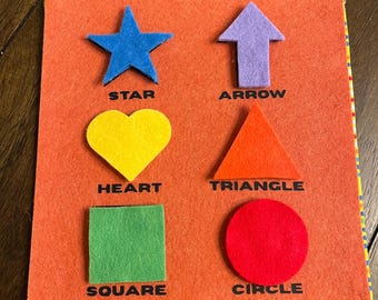 May include: An orange felt board with six colorful felt shapes: a blue star, a yellow heart, a green square, a red circle, a purple arrow, and an orange triangle. Each shape has its name printed below it in black.