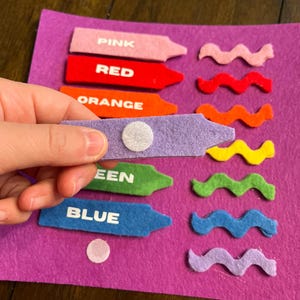 May include: A felt color matching activity set with crayon-shaped pieces labeled PINK, RED, ORANGE, GREEN, and BLUE. Each crayon has a matching wavy felt piece. The set is on a purple felt background.