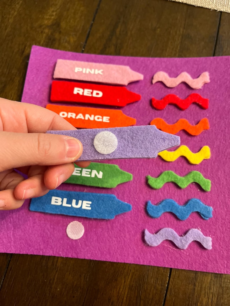 May include: A felt color matching activity set with crayon-shaped pieces labeled PINK, RED, ORANGE, GREEN, and BLUE. Each crayon has a matching wavy felt piece. The set is on a purple felt background.