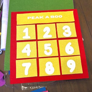 May include: A rectangular red felt board featuring a yellow felt header with the white text PEAK A BOO. Below the header are nine yellow felt squares arranged in a grid, each displaying a white felt number from one through nine.