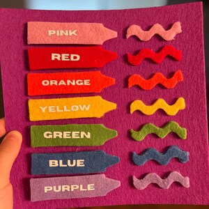 May include: A felt color learning board with seven crayon-shaped pieces labeled "PINK", "RED", "ORANGE", "YELLOW", "GREEN", "BLUE", and "PURPLE". Each crayon has a matching wavy felt shape. The background is a solid purple.