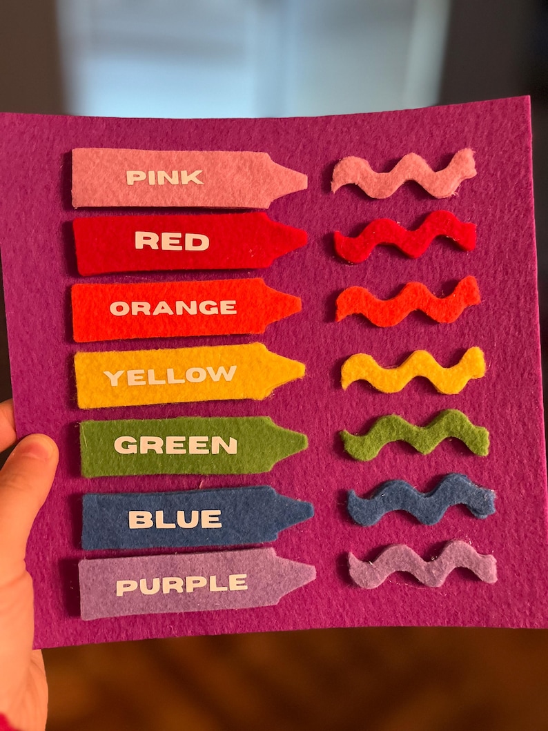 May include: A felt color learning board with seven crayon-shaped pieces labeled "PINK", "RED", "ORANGE", "YELLOW", "GREEN", "BLUE", and "PURPLE". Each crayon has a matching wavy felt shape. The background is a solid purple.