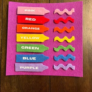 May include: A felt color learning set with pink, red, orange, yellow, green, blue, and purple crayons and wavy lines. Each crayon has the color name printed on it in white. The set is on a purple felt background.