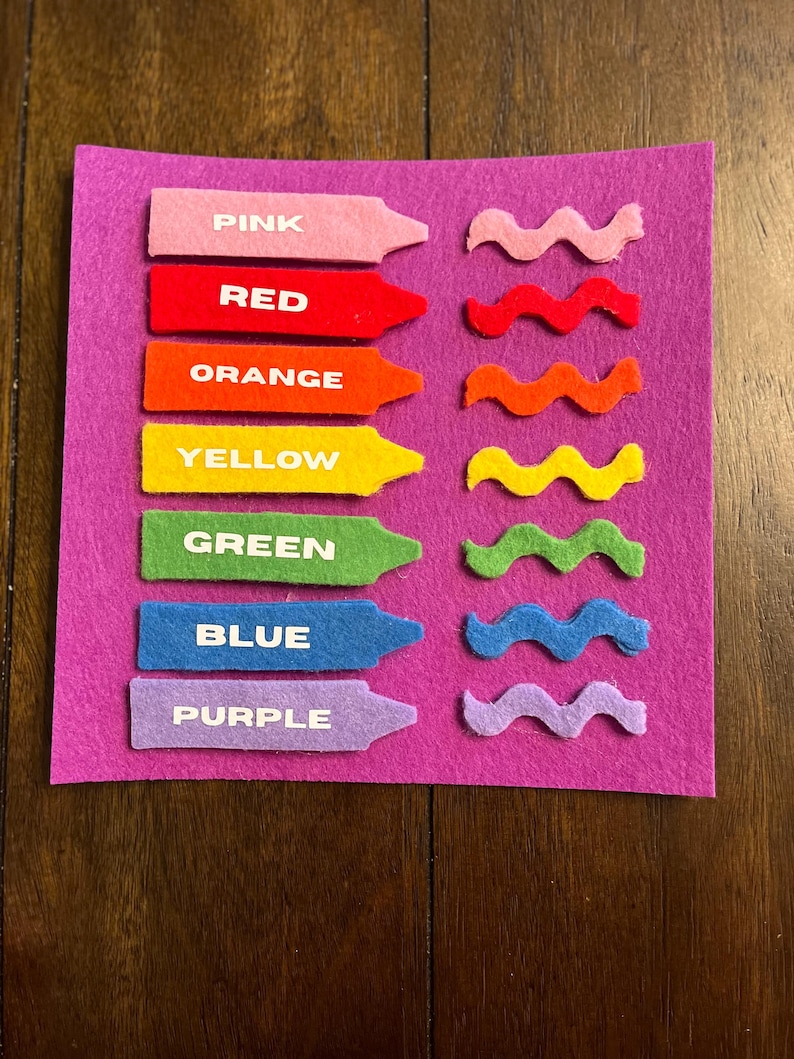May include: A felt color learning set with pink, red, orange, yellow, green, blue, and purple crayons and wavy lines. Each crayon has the color name printed on it in white. The set is on a purple felt background.