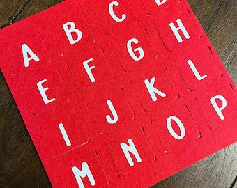 May include: A red felt alphabet puzzle with white uppercase letters. The letters A through P are visible, arranged in rows and columns. The puzzle is on a dark wooden surface.
