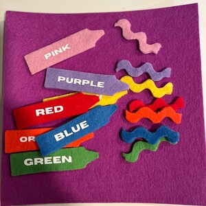 May include: A felt color learning set with crayon shapes and wavy lines on a purple background. The crayons are pink, purple, red, orange, blue, and green, each labeled with its color name in white. The wavy lines are pink, purple, yellow, orange, blue, and green.