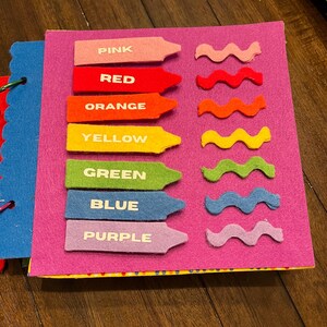 May include: A felt color learning book with a pink cover. The book features felt cutouts of crayons and wavy lines in pink, red, orange, yellow, green, blue, and purple. Each crayon has a color name printed on it.