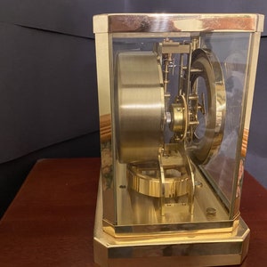 K&ouml;nnte beinhalten: Eine goldfarbene Uhr mit einem Glasgeh&auml;use, das die komplizierten inneren Mechanismen des Uhrwerks freilegt. Die Uhr steht auf einer Holzoberfl&auml;che.