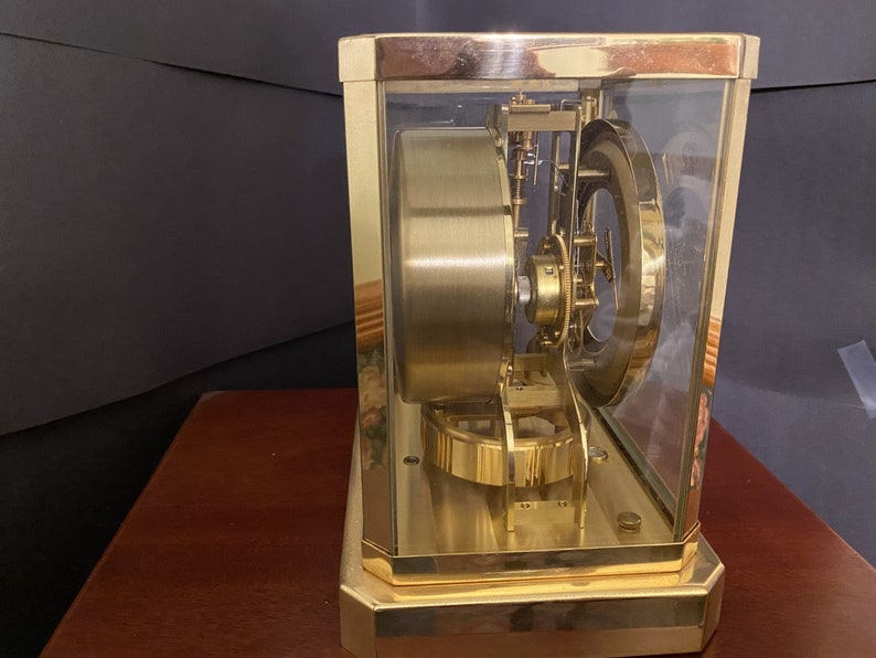 K&ouml;nnte beinhalten: Eine goldfarbene Uhr mit einem Glasgeh&auml;use, das die komplizierten inneren Mechanismen des Uhrwerks freilegt. Die Uhr steht auf einer Holzoberfl&auml;che.