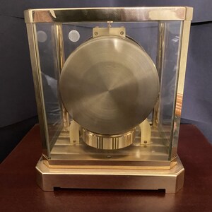 K&ouml;nnte beinhalten: Eine goldfarbene Uhr mit einem Glasgeh&auml;use und einem runden, goldfarbenen Zifferblatt. Die Uhr steht auf einer Holzoberfl&auml;che.