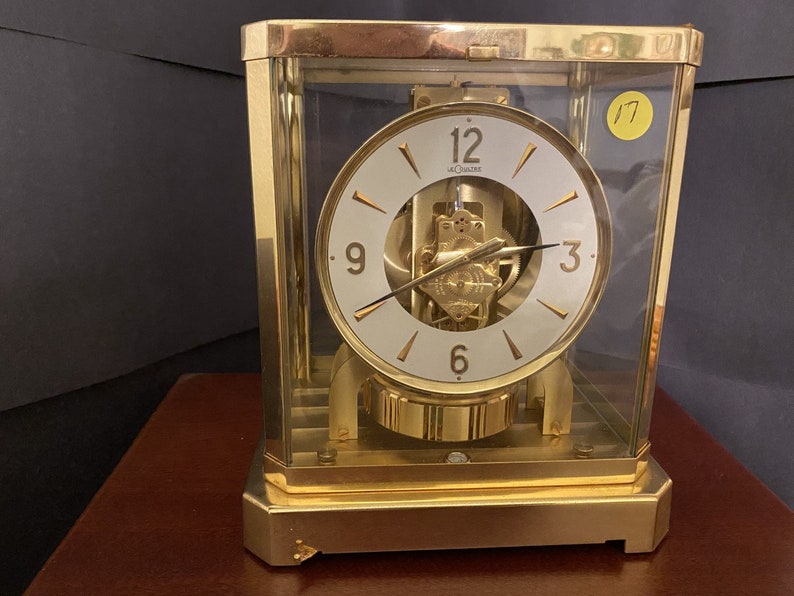 K&ouml;nnte beinhalten: Eine goldfarbene, quadratische Uhr mit einem wei&szlig;en Zifferblatt und schwarzen r&ouml;mischen Ziffern. Die Uhr ist in Glas gefasst und hat ein sichtbares Uhrwerk. Das Zifferblatt der Uhr zeigt oben "12", rechts "3", unten "6" und links "9". Das Zifferblatt der Uhr enth&auml;lt auch den Text "LeCoultre".