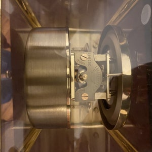 K&ouml;nnte beinhalten: Nahaufnahme eines goldfarbenen Uhrwerks mit einer durchsichtigen Glasabdeckung. Das Uhrwerk hat ein rundes Zifferblatt mit Markierungen und einen kleinen, runden Knopf, auf dem die Nummer "296892" aufgedruckt ist. Der Knopf ist mit "RIMOS" beschriftet.