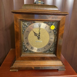 May include: A wooden mantel clock with a gold face and Roman numeral hour markers. The clock has a gold trim and a small label that reads "Westclox".