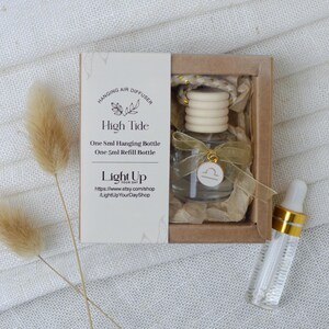 May include: A brown cardboard box with a clear glass bottle hanging from a gold and white string. The bottle has a wooden stopper and a gold charm with the Libra zodiac symbol. The box has a white label with the text "High Tide" and "One 5ml Hanging Bottle, One 5ml Refill Bottle". The label also includes the text "Light Up Your Day" and a website address. A clear glass bottle with a gold cap is sitting next to the box.