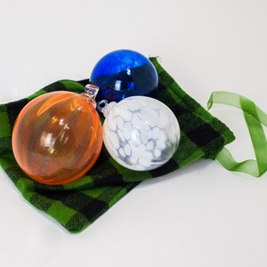 May include: Three glass ornaments in a green and black plaid bag with a green ribbon. The ornaments are orange, white with white swirls, and blue.