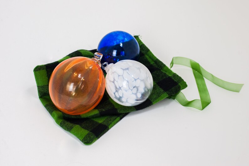 May include: Three glass ornaments in a green and black plaid bag with a green ribbon. The ornaments are orange, white with white swirls, and blue.