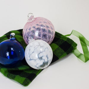 May include: Three glass ornaments in a green and black plaid bag with a green ribbon. The ornaments are blue, white with gray swirls, and pink with purple swirls.